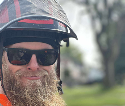 Firefighter with a helmet and sunglasses, close-up shot smiling with a beard against a blurred outdoor background.
