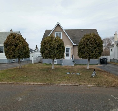 Front view of a beige house with a steep gable roof, three large trimmed trees in the lawn, and a driveway on the right.